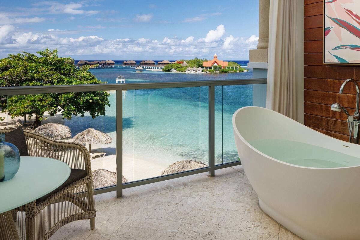 Une salle de bain moderne avec une baignoire sur pieds, un balcon vitré offrant une vue sur une plage tropicale et des bungalows sur pilotis.
