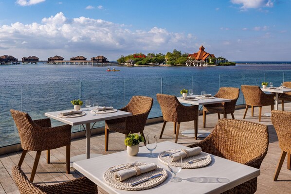 A seaside restaurant with tables and wicker chairs, overlooking the sea and a distant island with overwater bungalows.