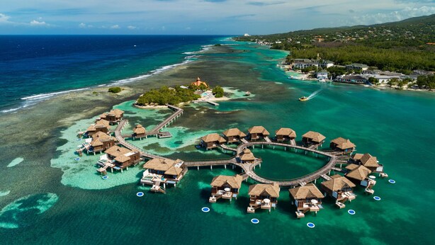 Vista aérea de bungalows sobre el agua conectados por pasarelas, rodeados de aguas turquesas cristalinas y una costa distante.