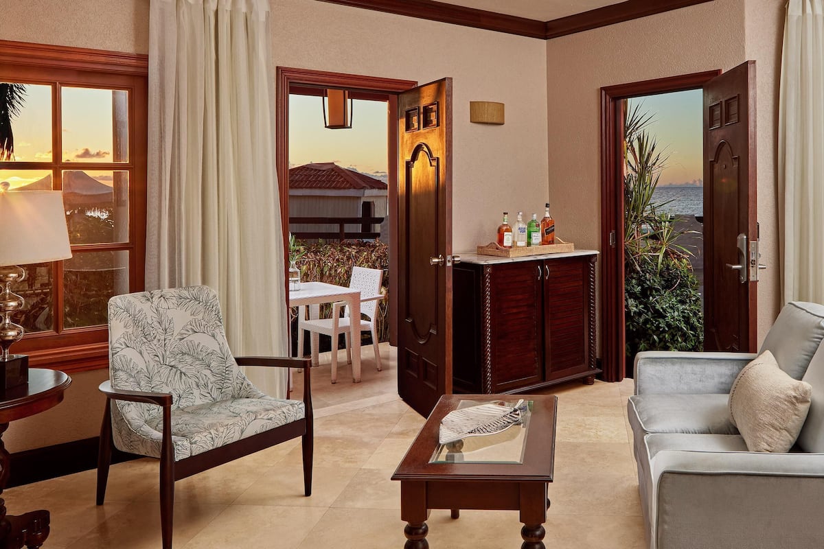 Un salon comprenant un canapé, un fauteuil et une table avec une lampe. Une porte donne accès à un balcon d’où l’on peut admirer une montagne et un plan d’eau.
