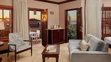 Una sala de estar con sofá, sillón y una mesa con lámpara. Hay una puerta que da a un balcón con vistas a una montaña y una masa de agua.