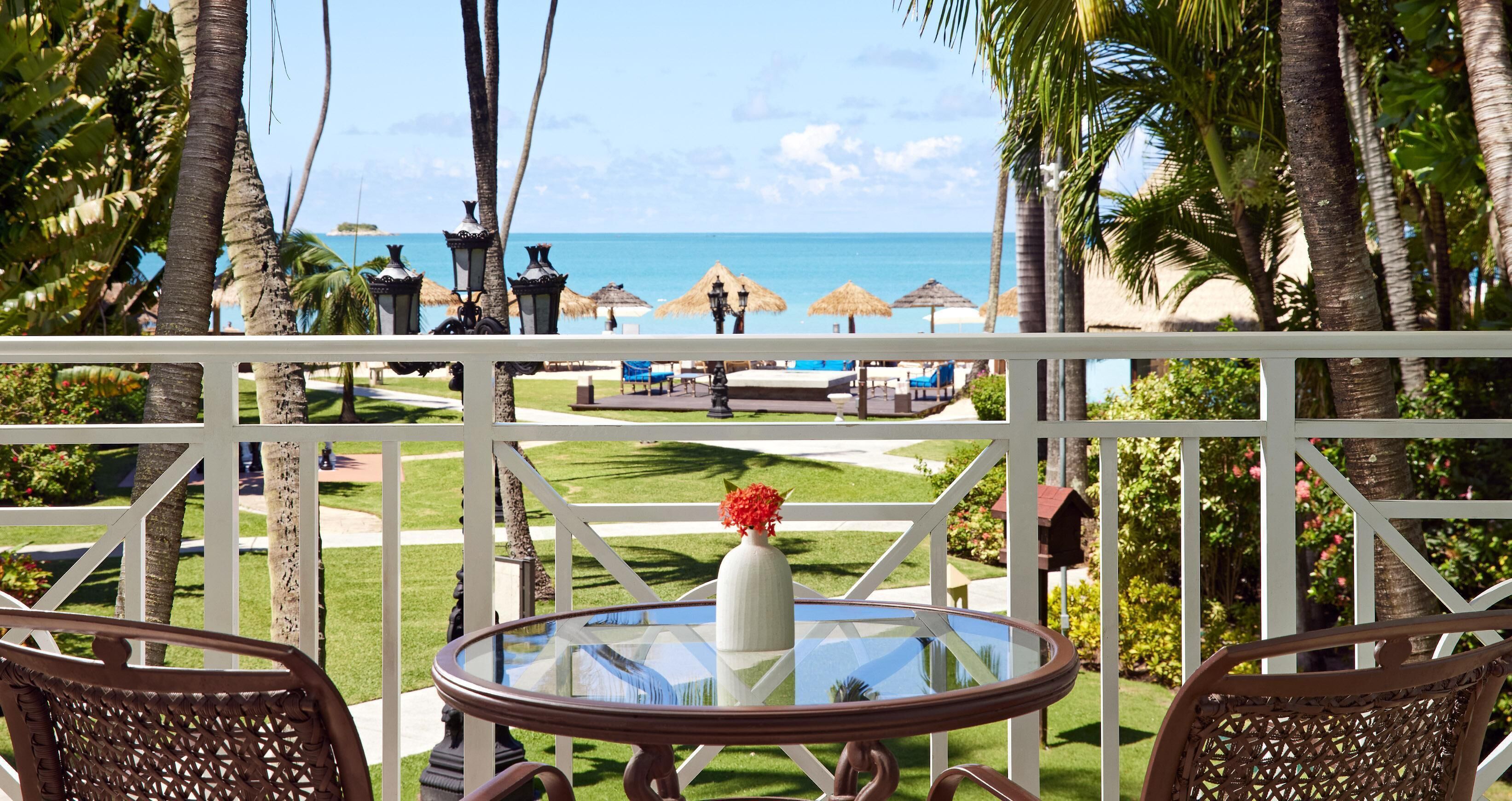A glass-top table with a white vase and a wicker chair in a tropical setting with a view of the beach and palm trees.