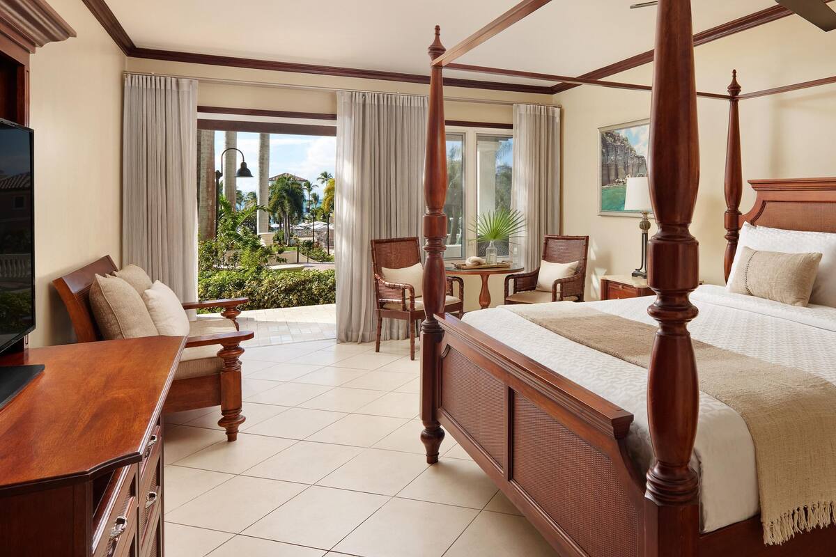 Un lit à baldaquin avec une tête de lit en bois, un bureau en bois doté de tiroirs, une télévision et une vue sur un jardin tropical grâce à de grandes fenêtres.