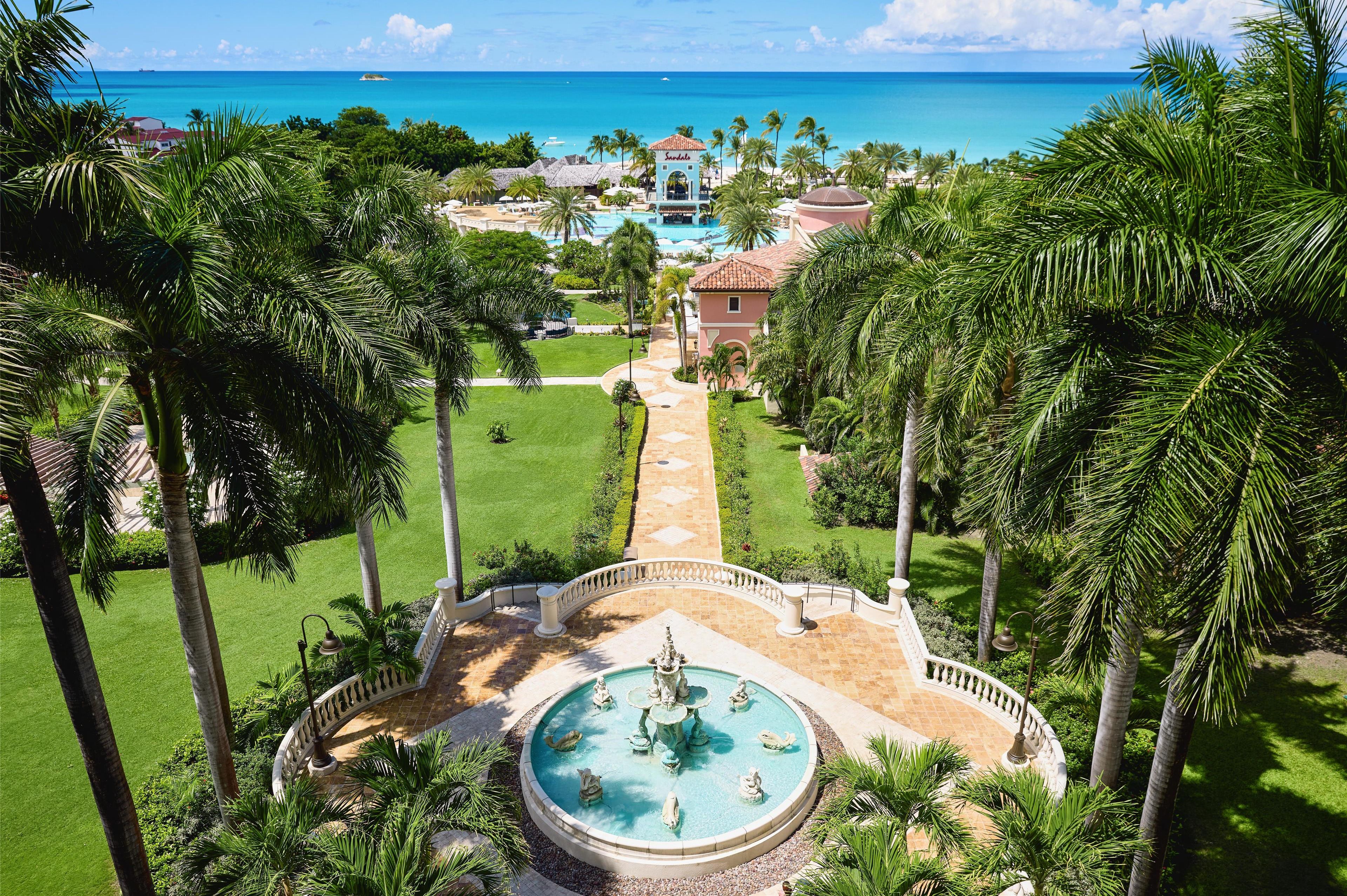 A resort with a central fountain, palm trees, and a view of the ocean.