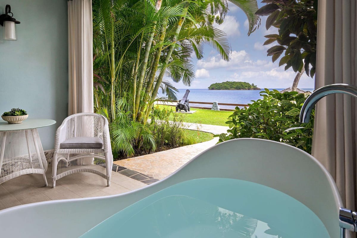 A bathroom with a bathtub, wicker chairs, a small table with a plant, and a view of a tropical landscape with a body of water and greenery.