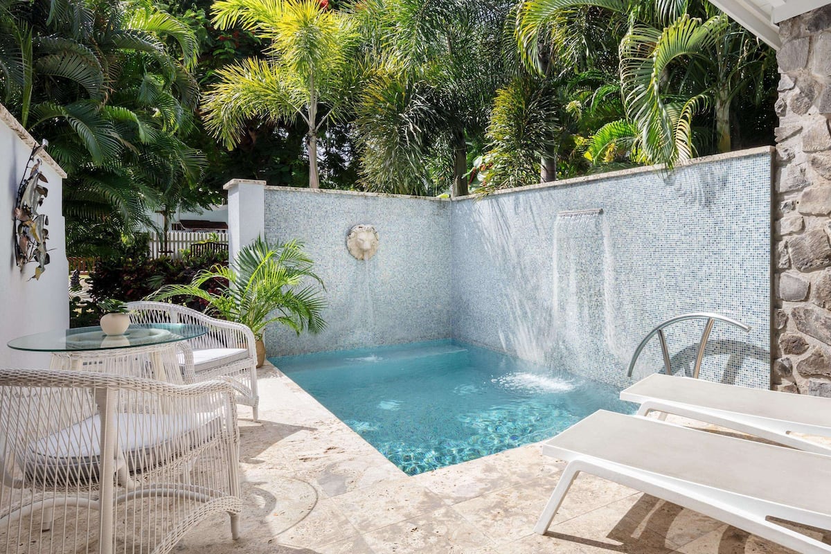 A private pool with a stone wall and glass table, surrounded by lounge chairs and tropical plants.