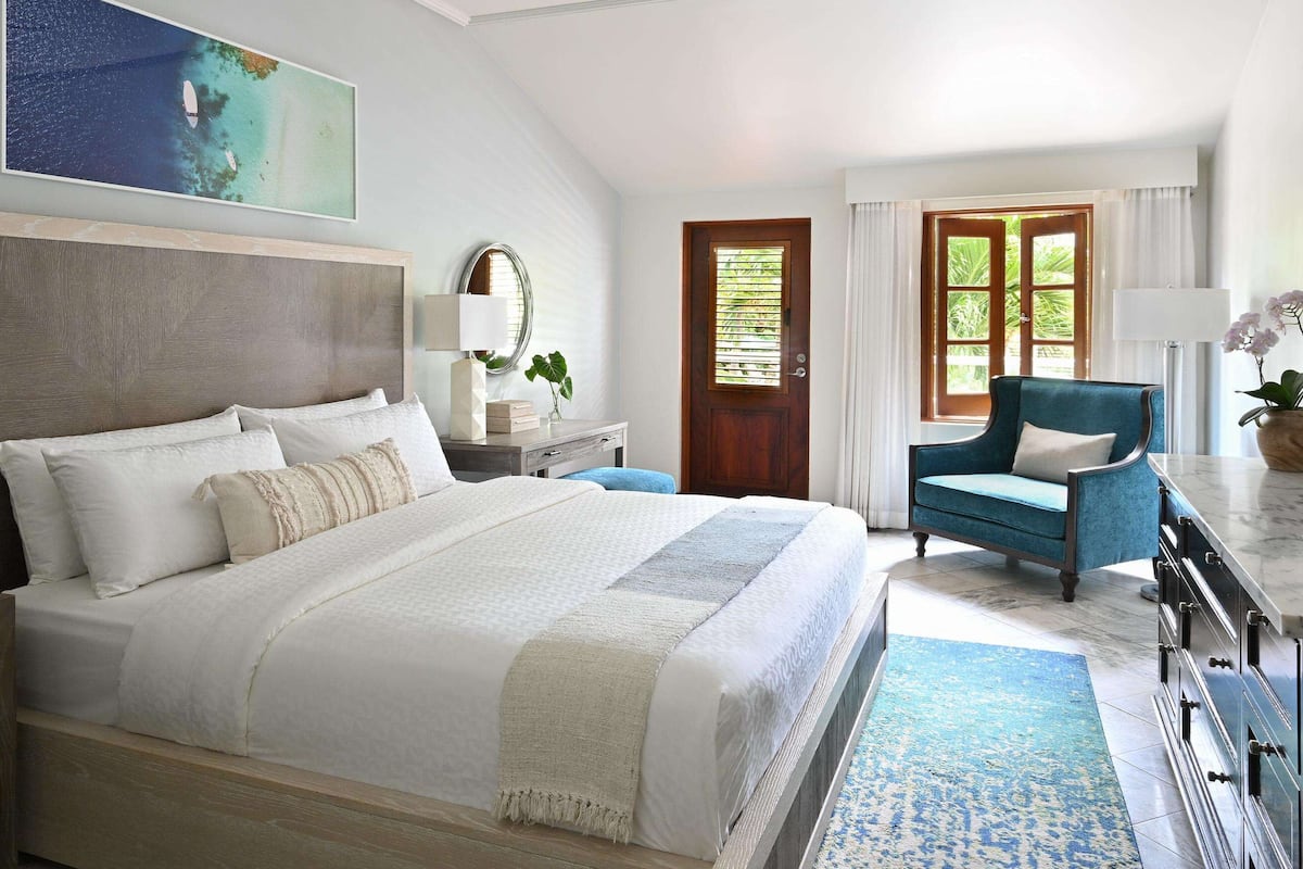 A spacious bedroom with a large bed, a wooden nightstand, a desk, a blue armchair, and a television.