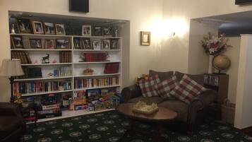 A cozy living room with a bookshelf filled with books and framed photos, a patterned carpet, a brown leather sofa with plaid cushions, and a small wooden coffee table.