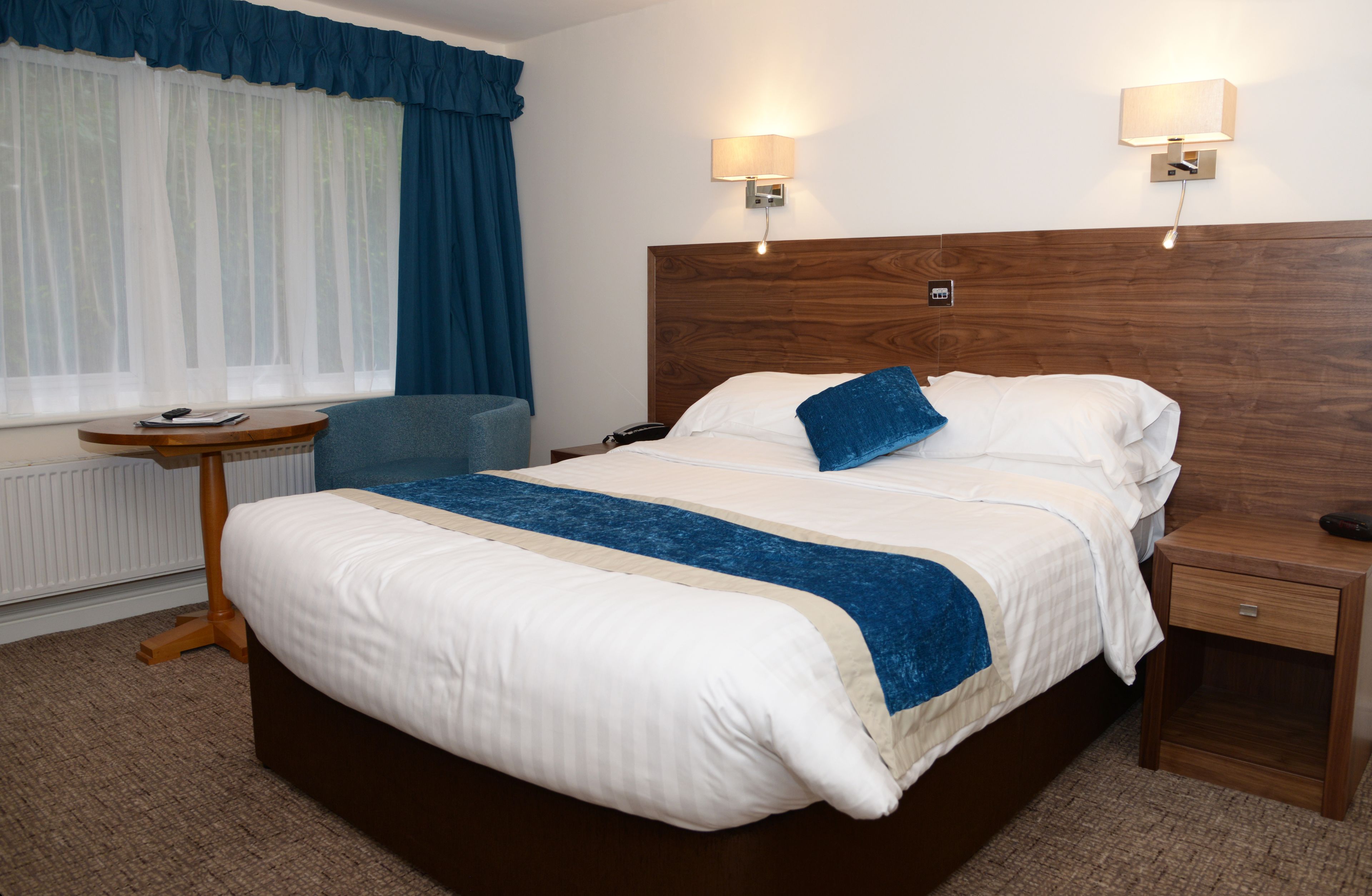 Una cama bien hecha con un cojín azul decorativo, una mesita de noche con lámpara y una silla en una habitación de hotel.