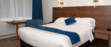 Una cama bien hecha con un cojín azul decorativo, una mesita de noche con lámpara y una silla en una habitación de hotel.