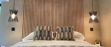 Un dormitorio moderno con cama, dos almohadones estampados, una colcha con diseño, un cabecero de listones de madera y dos lámparas colgantes.