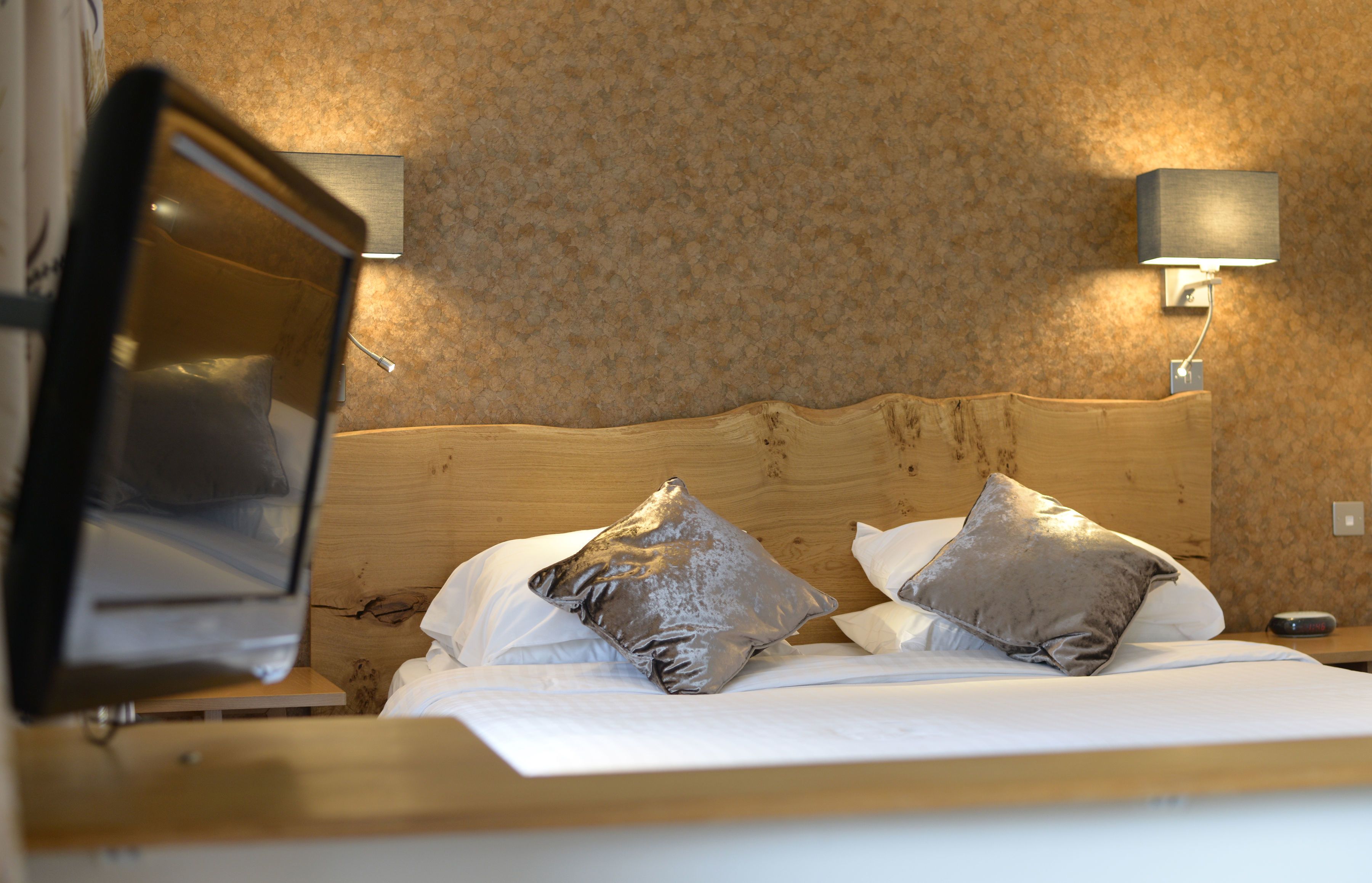 A hotel room with a wooden headboard, two pillows, a bedside table, and a wall-mounted lamp.