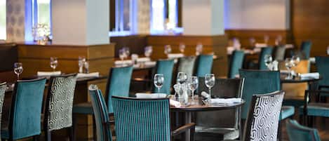 A restaurant interior with teal patterned chairs, dark wood tables, and hanging pendant lights.