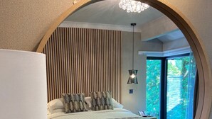 A bedroom with a circular mirror reflecting a bed with patterned bedding, a bedside table with a lamp, and a view of greenery outside. - Waterloo Hotel Lodge (Betws-Y-Coed)