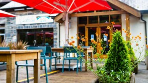 An outdoor seating area with wooden tables and chairs, red umbrellas, and a building with a stone facade in the background. - Waterloo Hotel Lodge (Betws-Y-Coed)