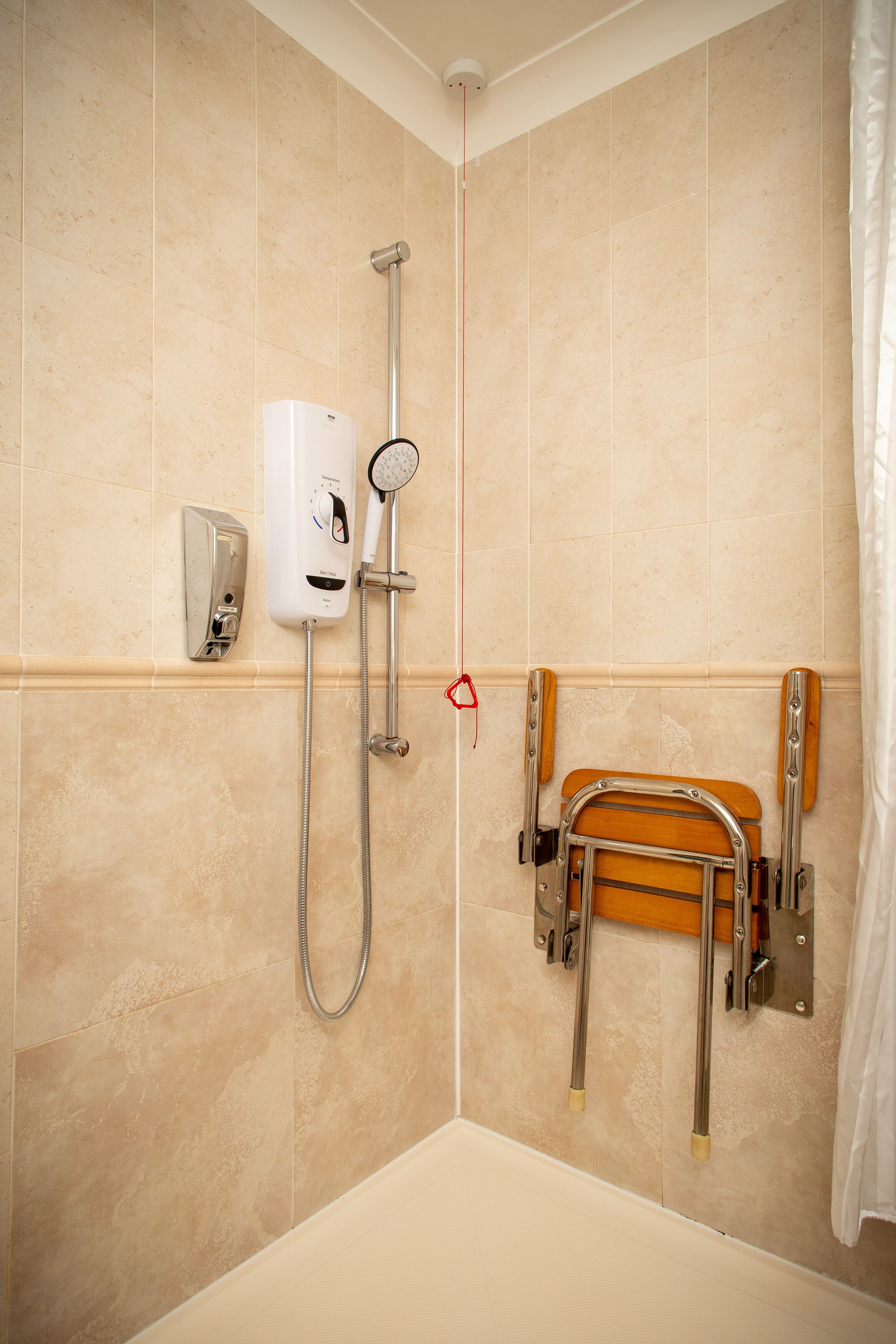A bathroom with beige tiles, a shower with a handheld showerhead, a handheld shower attachment, a soap dispenser, and a wooden bath seat.