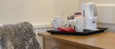 A chair with a patterned fabric, a wooden table with a tray holding a coffee maker, cups, and a red bag.