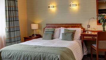 Une chambre d’hôtel avec un grand lit, des tables de chevet, un bureau et une chaise. Il y a deux lampes fixées au mur et un miroir au-dessus du bureau.