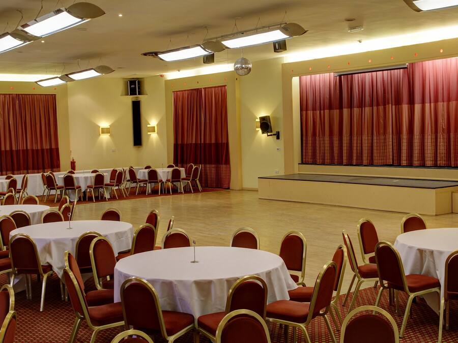 Un salón de conferencias con varias mesas redondas y sillas rojas, equipado con luces en el techo y un escenario con cortinas.