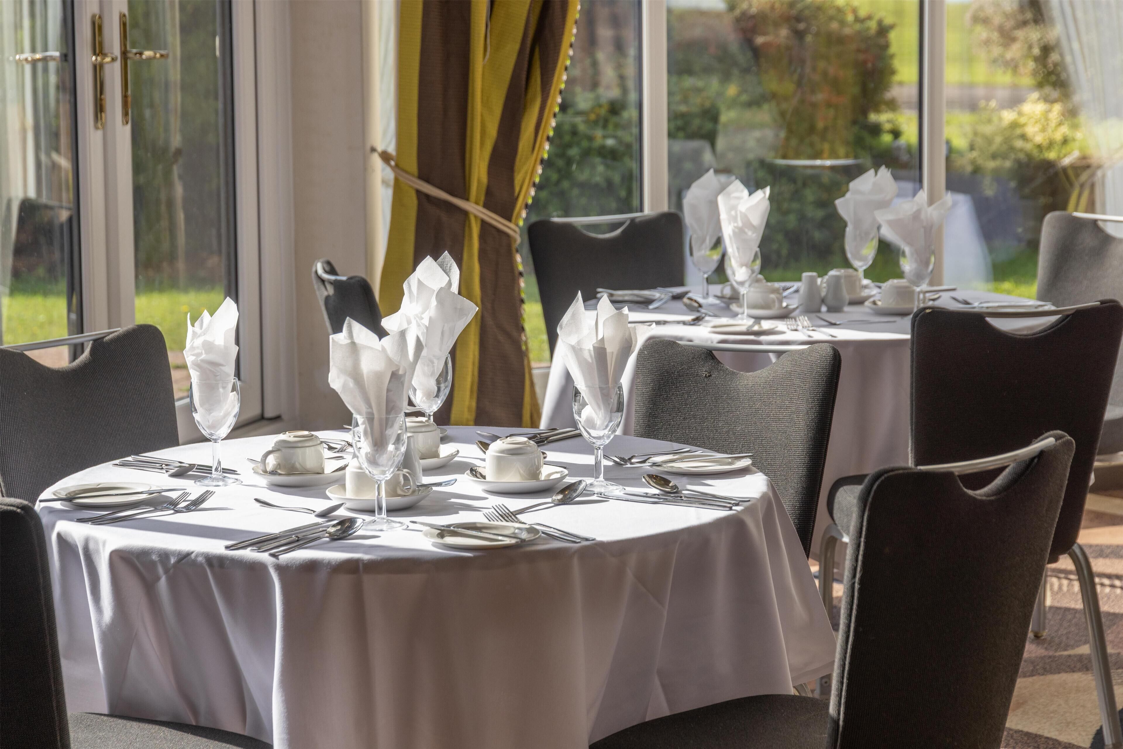 A dining area with round tables set for four, each with white tablecloths, neatly arranged cutlery, and folded napkins.