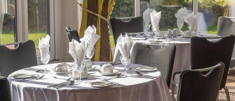 A dining area with round tables set for four, each with white tablecloths, neatly arranged cutlery, and folded napkins.