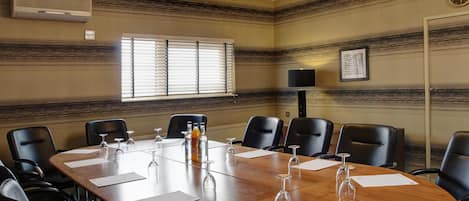 Un salón de conferencias con una gran mesa de madera, sillas de cuero negro, un candelabro y una ventana con persianas.