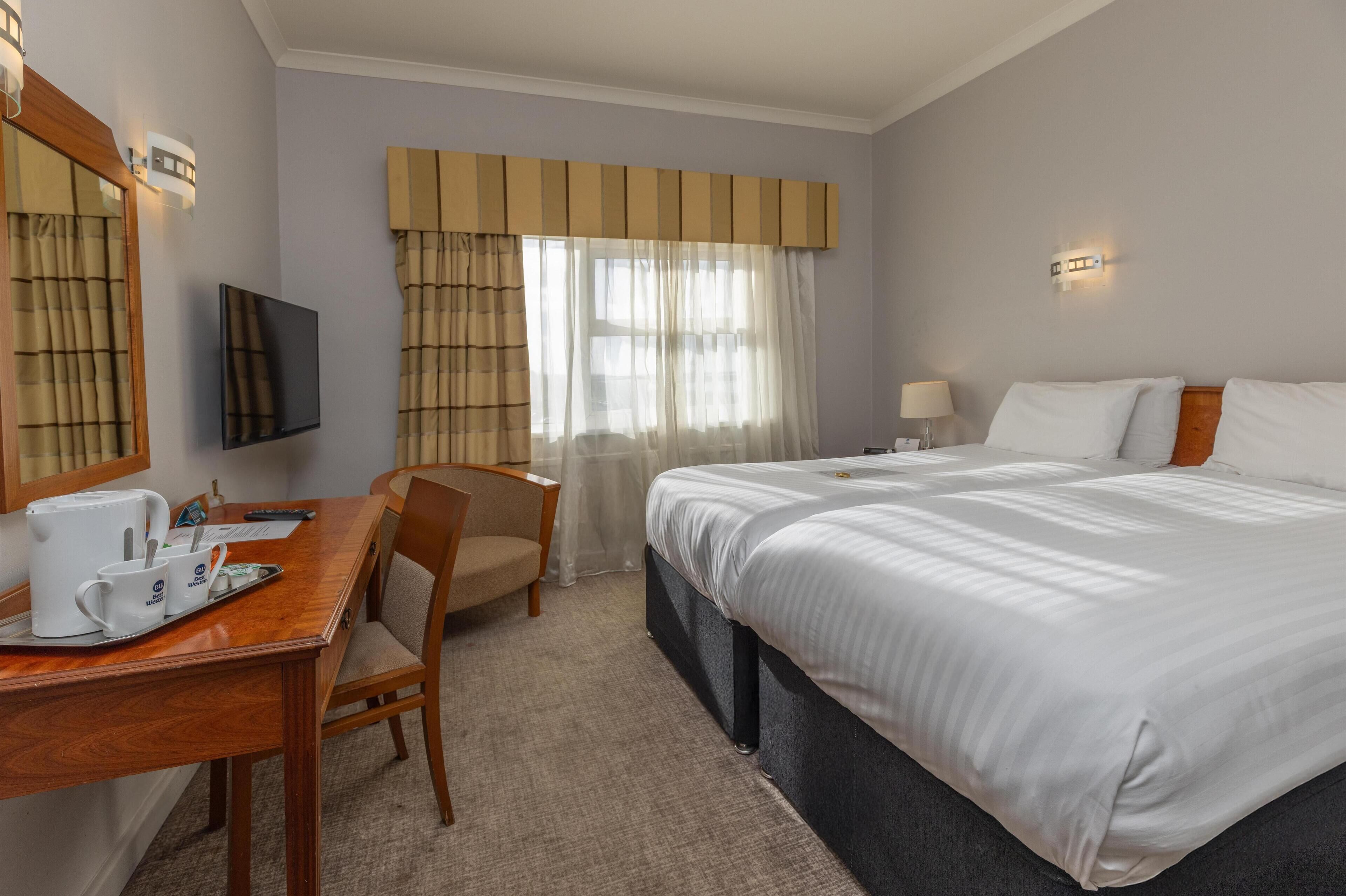Ein Hotelzimmer mit einem großen Bett, einem Schreibtisch mit Kaffeemaschine, einem Sessel, einem Fernseher und einem Vorhangfenster.