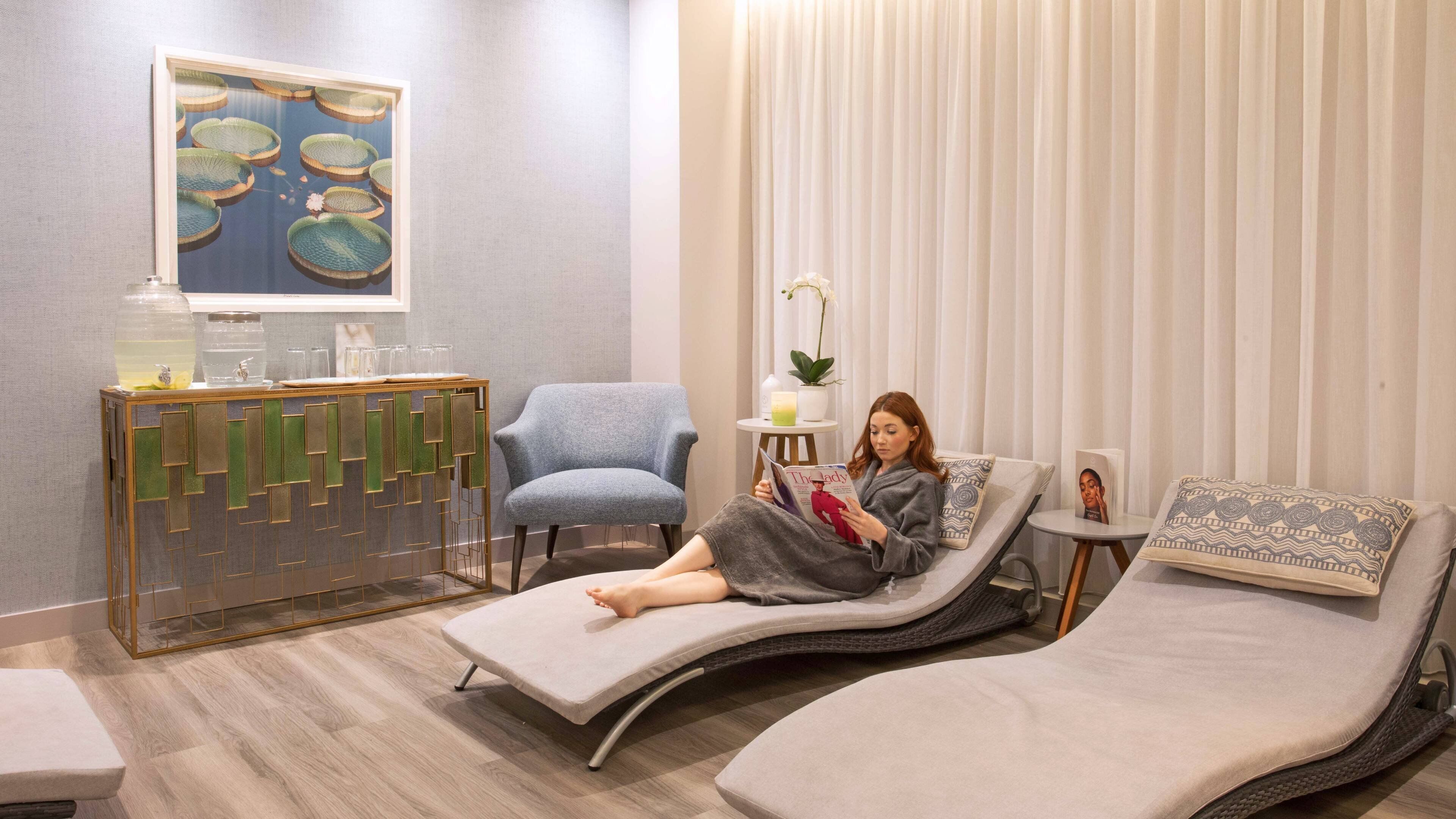Une salle de spa comprenant une table de massage, un fauteuil bleu, une petite table d’appoint avec un magazine et un élément décoratif mural.