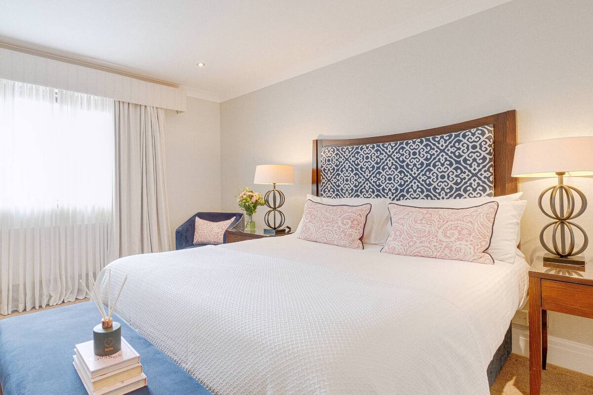 A hotel room with a large bed, a nightstand with a lamp, a chair, and a window with curtains.
