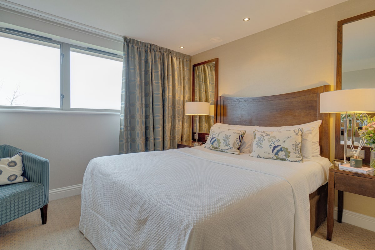 A hotel room with a large bed, a chair, a nightstand with a lamp, and a window with curtains.