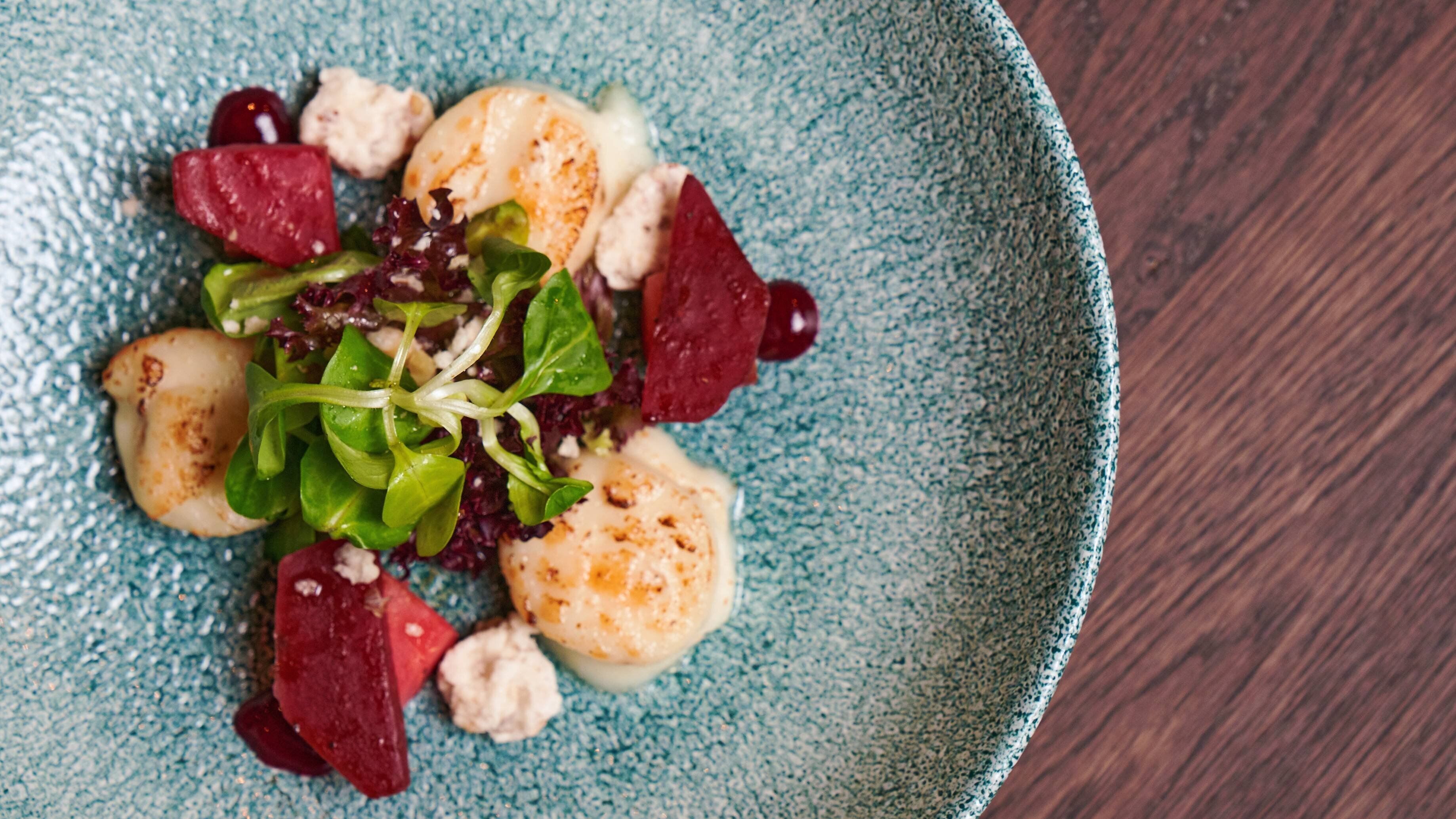 A plate of food with scallops, greens, and beetroot on a textured blue plate.