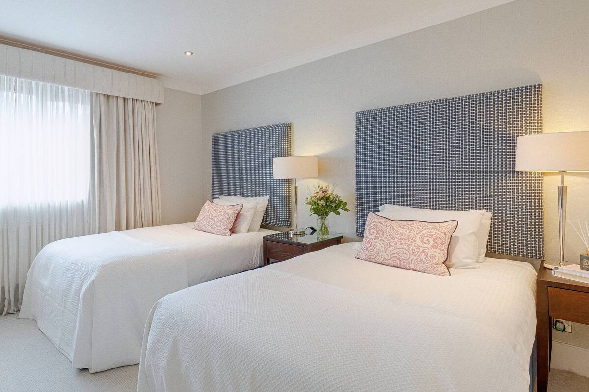 A hotel room with two beds, a large window with sheer curtains, a nightstand with a lamp, and a patterned headboard.