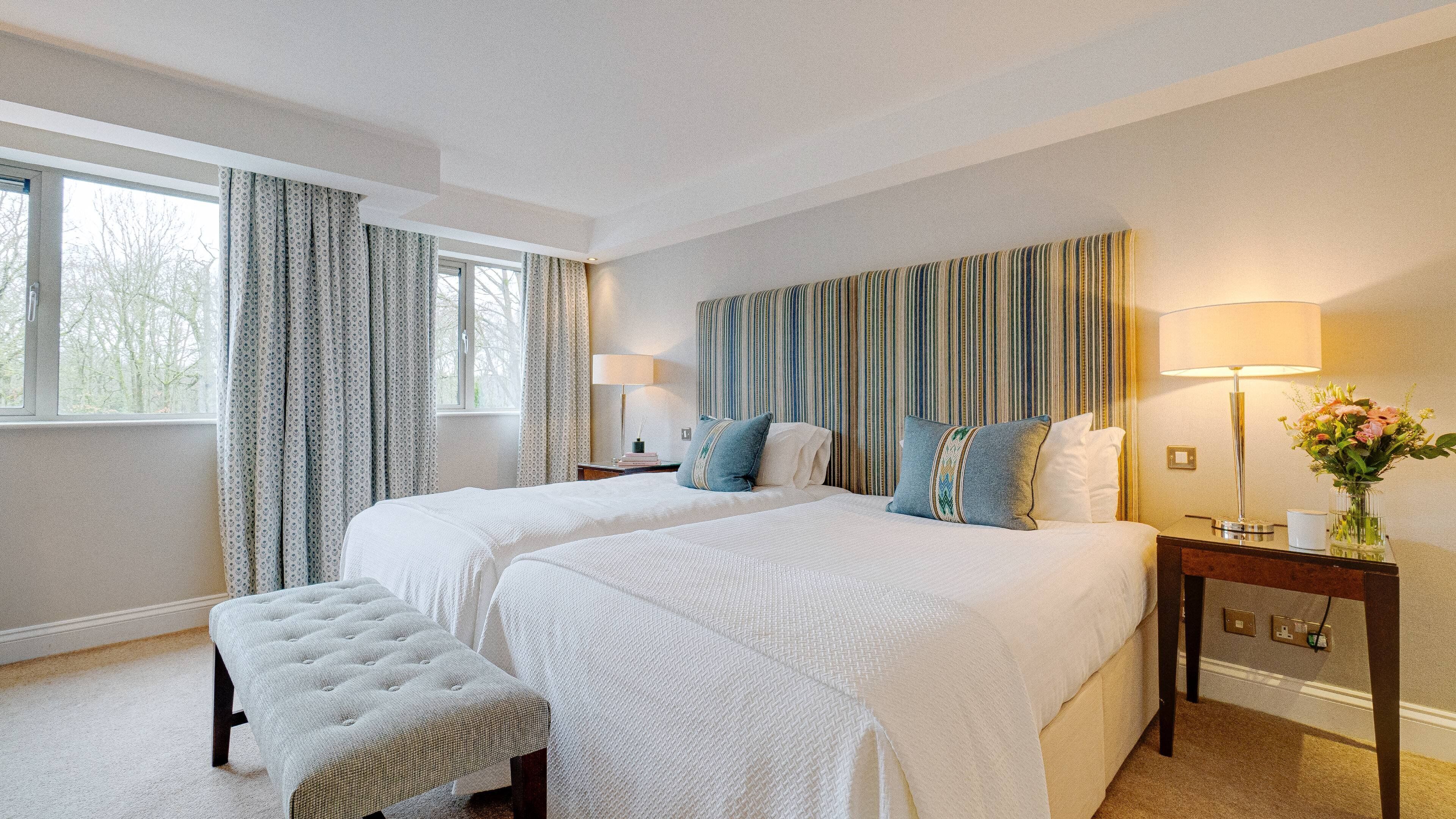 Habitación de hotel con dos camas, un ventanal con cortinas, una mesita de noche con lámpara y una pequeña mesa con un jarrón de flores.