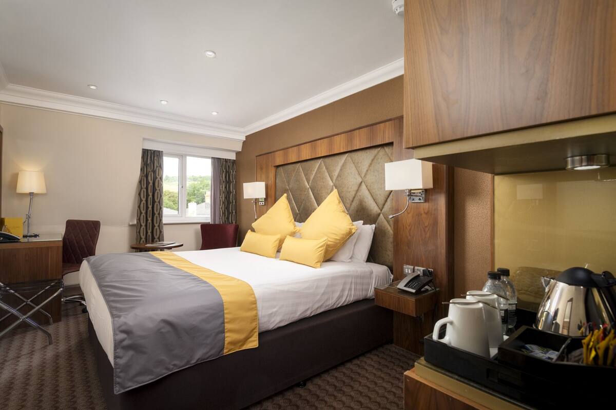 A hotel room with a large bed, a desk, a chair, a desk lamp, a telephone, a coffee maker, and a mirror.