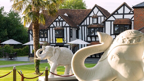 A traditional Tudor-style building with a white and black timber framing exterior, a brick chimney, and a surrounding garden with palm trees and outdoor seating. - Donnington Manor Hotel (Sevenoaks)