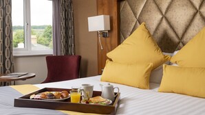 A hotel room with a bed, a bedside table, a chair, a window with curtains, and a view of greenery outside. - Donnington Manor Hotel (Sevenoaks)