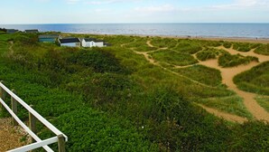 Paesaggio costiero con spiaggia sabbiosa, dune erbose e alcuni piccoli edifici.