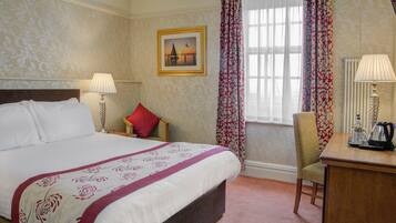 A hotel room with a large bed, two bedside lamps, a chair, a desk with a kettle, and a window with curtains.