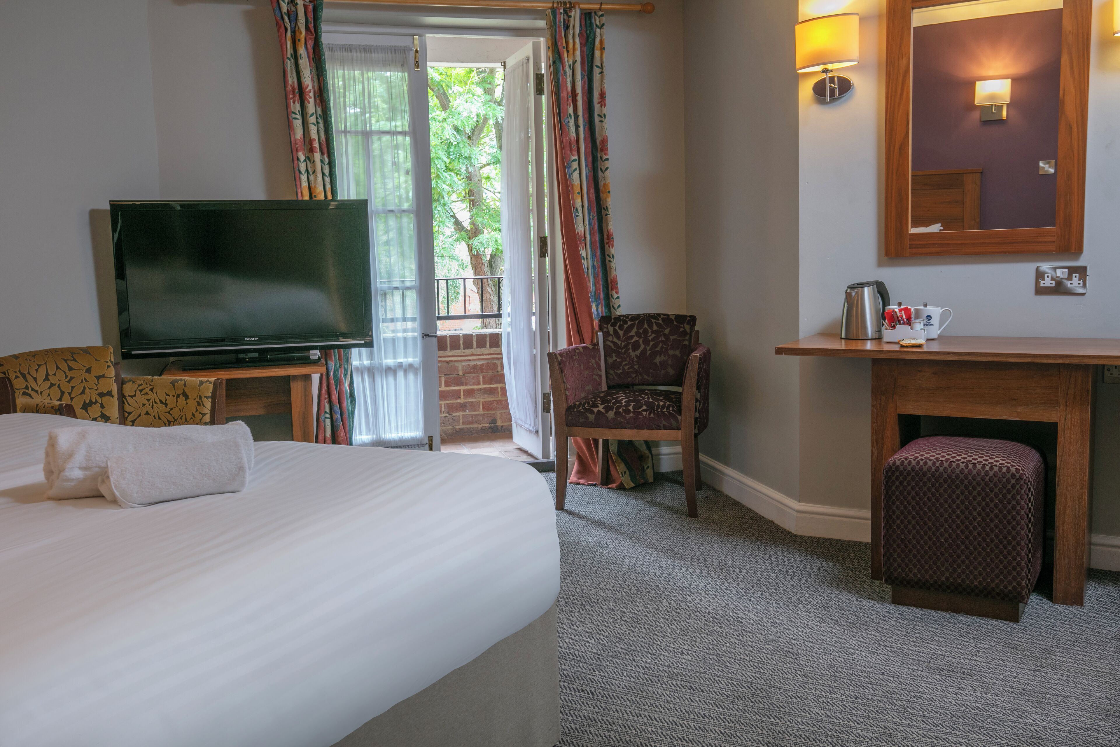 Habitación de hotel con cama, televisor, escritorio con tetera, silla y una pequeña otomana.