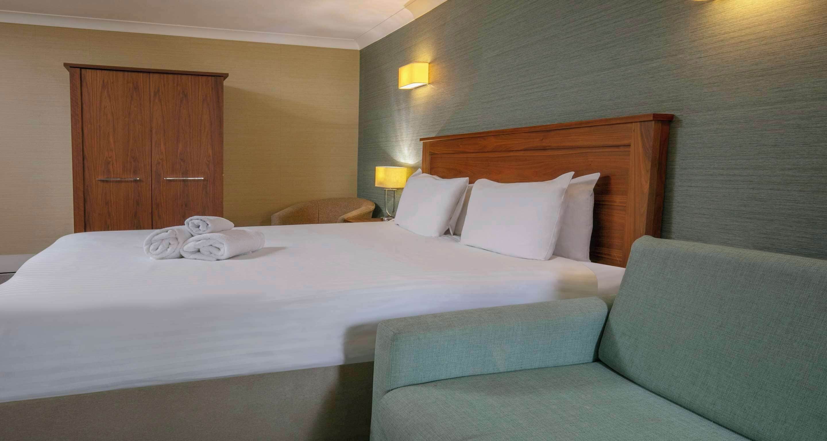 A hotel room with a large bed, a wooden wardrobe, a green armchair, and two bedside lamps.