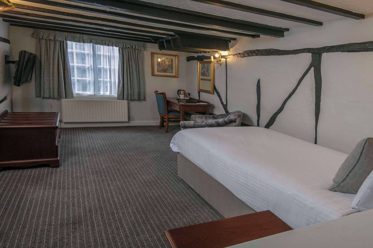 A bedroom with a bed, a desk, and a chair. There is a window with curtains and a radiator. The room has exposed wooden beams on the ceiling.