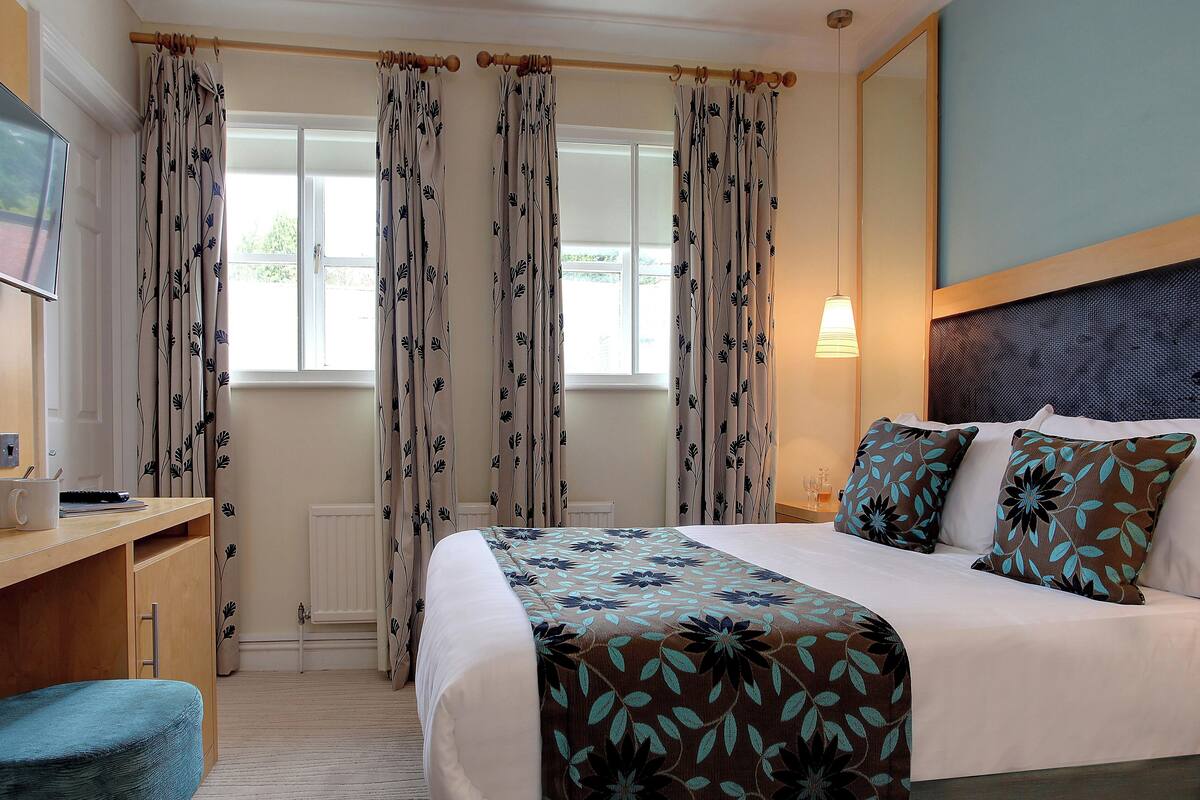 A hotel room with a bed, a desk, and a chair. There is a window with curtains, a bedside lamp, and a radiator.