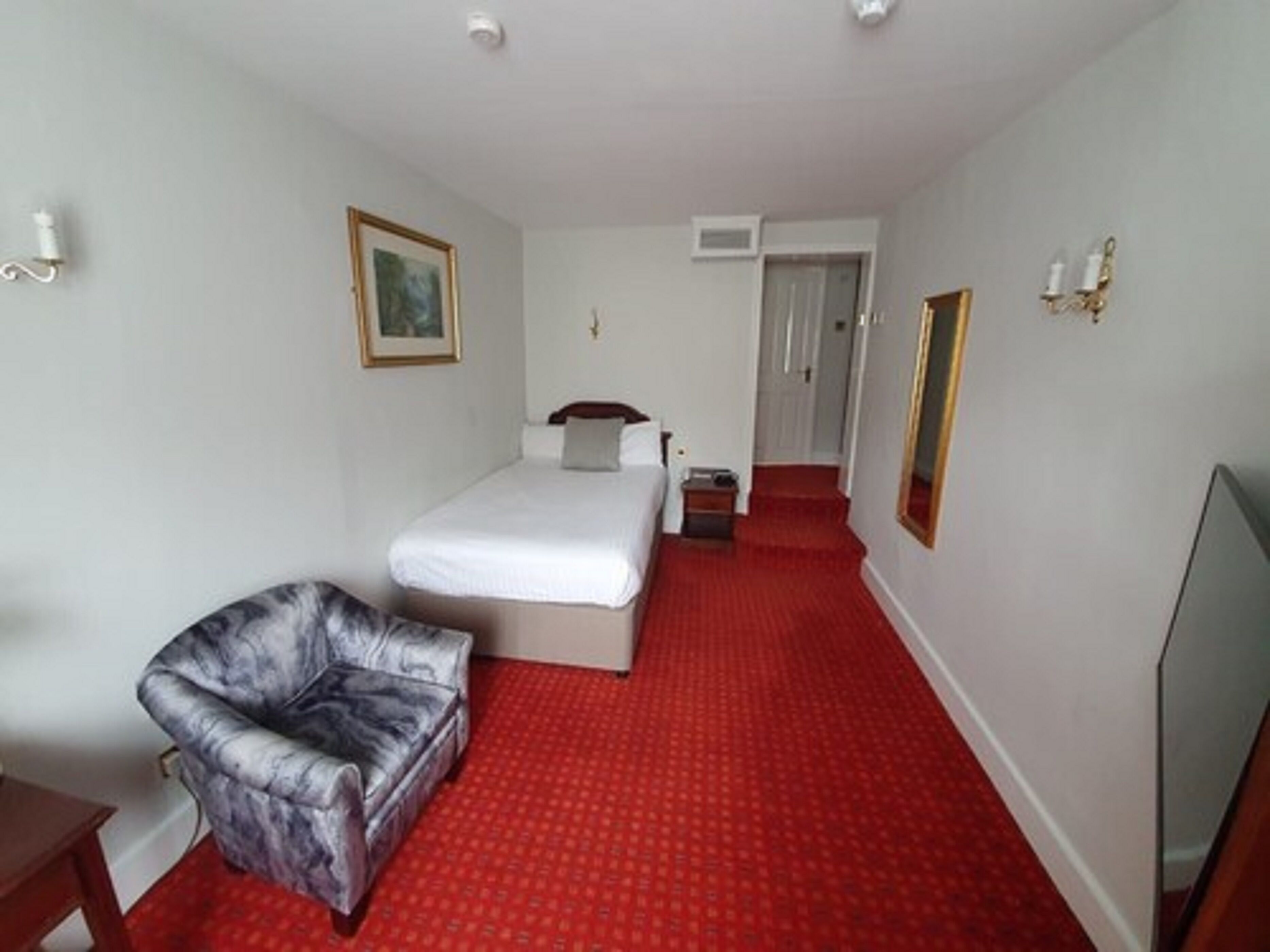 A hotel room with a bed, a chair, a small table, a mirror, and a red carpet.