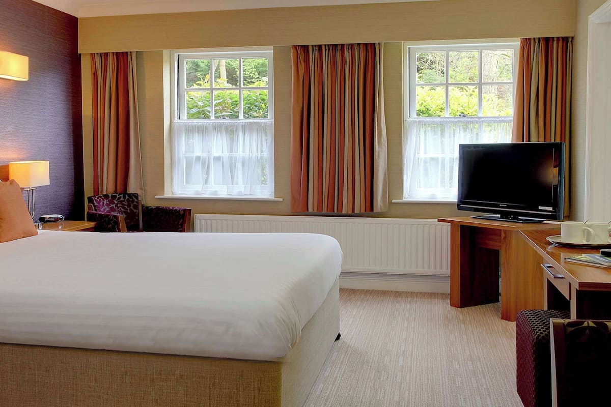 A hotel room with a large bed, a desk with a television, and windows with curtains.
