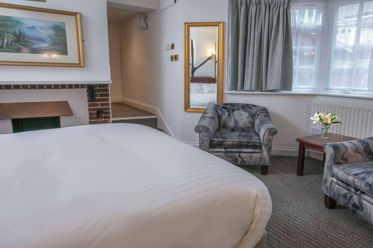 A hotel room with a large bed, two armchairs, a small table with a vase, a fireplace, a mirror, and a window with curtains.