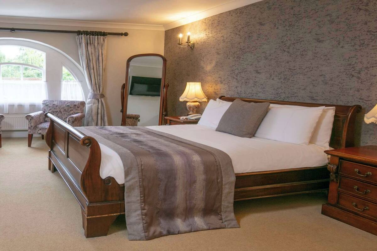 A bedroom with a large bed, two armchairs, a nightstand, a mirror, and a window with curtains.