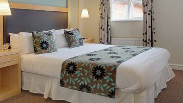 A neatly made bed with floral-patterned bedding, a bedside table with a lamp, and a window with curtains.
