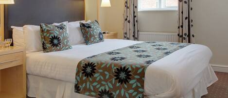 A neatly made bed with floral-patterned bedding, a bedside table with a lamp, and a window with curtains.