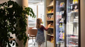 A person reaching for items in a refrigerated display case in a room with a plant, chairs, and a view outside.