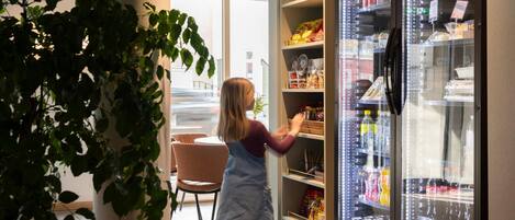 Una persona alcanzando productos en un refrigerador en una habitación con una planta, sillas y vistas al exterior.